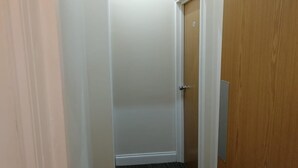 Un corridoio stretto con pavimento rivestito di moquette, una porta in legno con maniglia metallica e una porta bianca sullo sfondo.