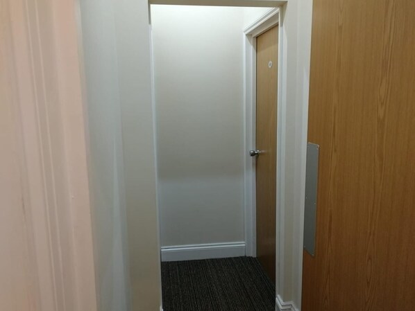 A narrow hallway with a carpeted floor, a wooden door with a metal handle, and a white door in the background. - Daisy Hotel Kings Cross (London)