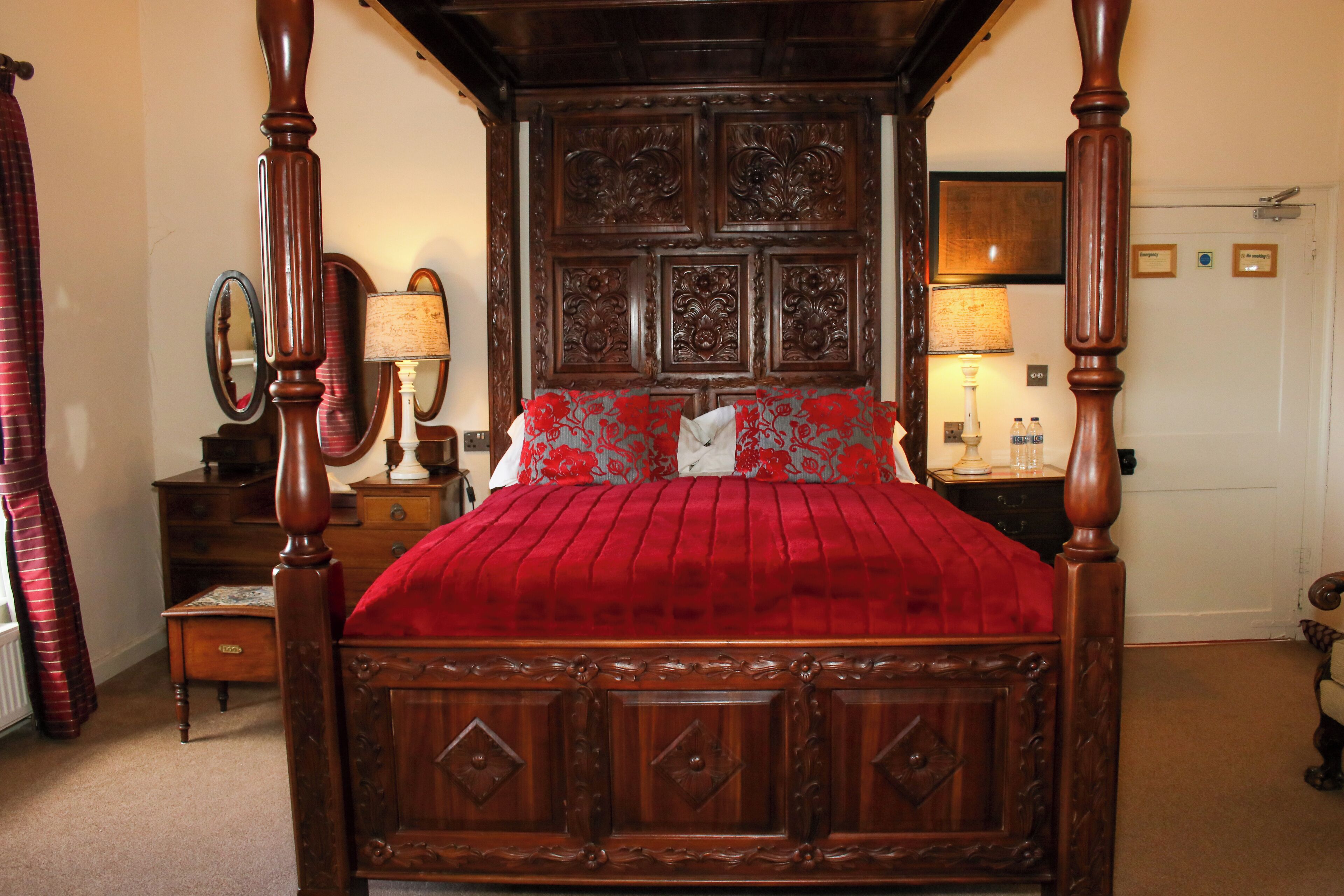 Una cama con dosel con un edredón rojo, un cabecero de madera con intrincados grabados y dos lámparas de noche.