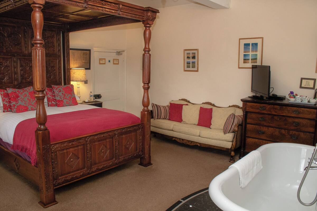 A four-poster bed with a red bedspread, a cream-colored sofa with red and white cushions, a wooden dresser with drawers, and a white clawfoot bathtub.