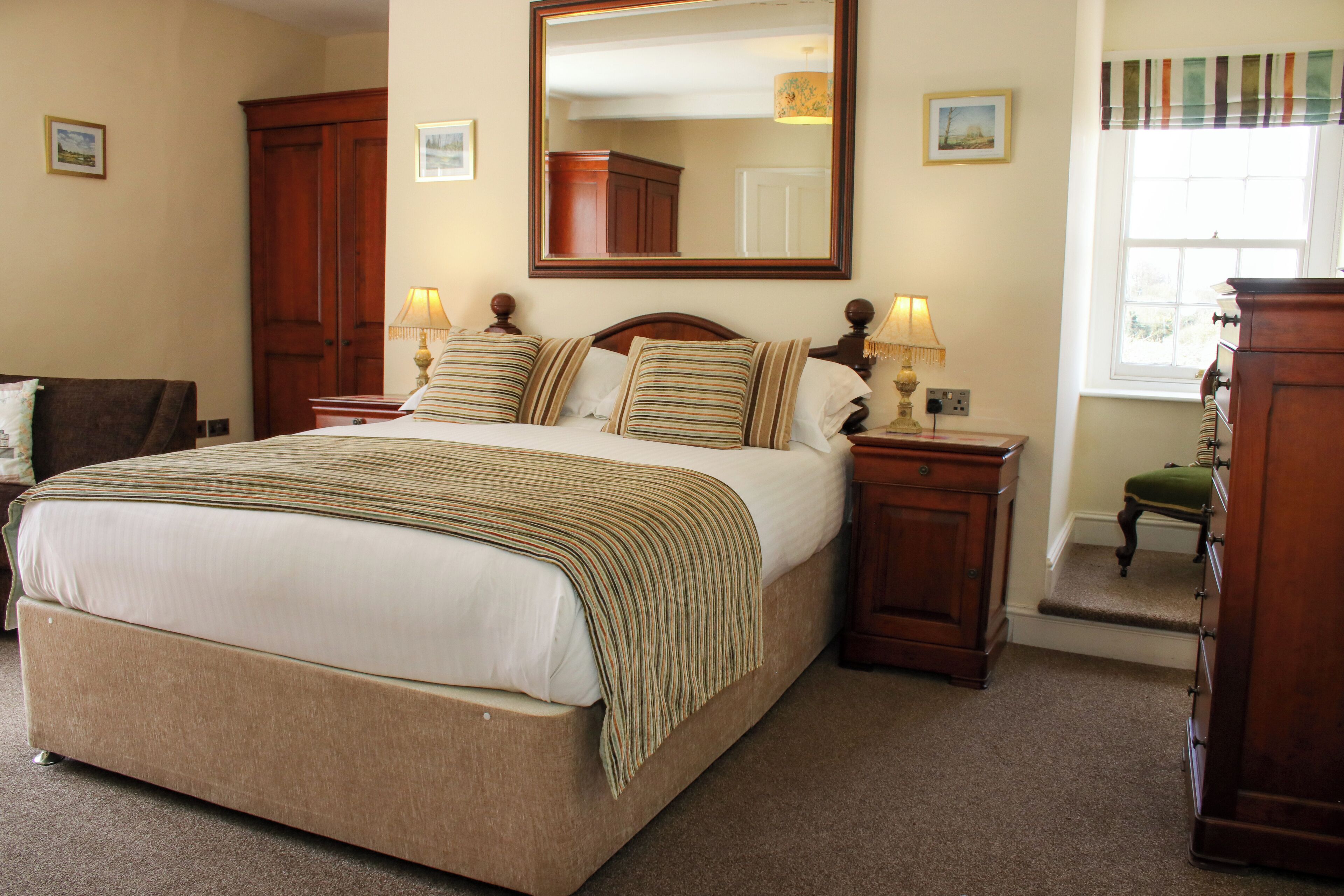 A bedroom with a large bed, wooden headboard, two bedside tables with lamps, a mirror above the bed, a wooden wardrobe, and a window with a view.