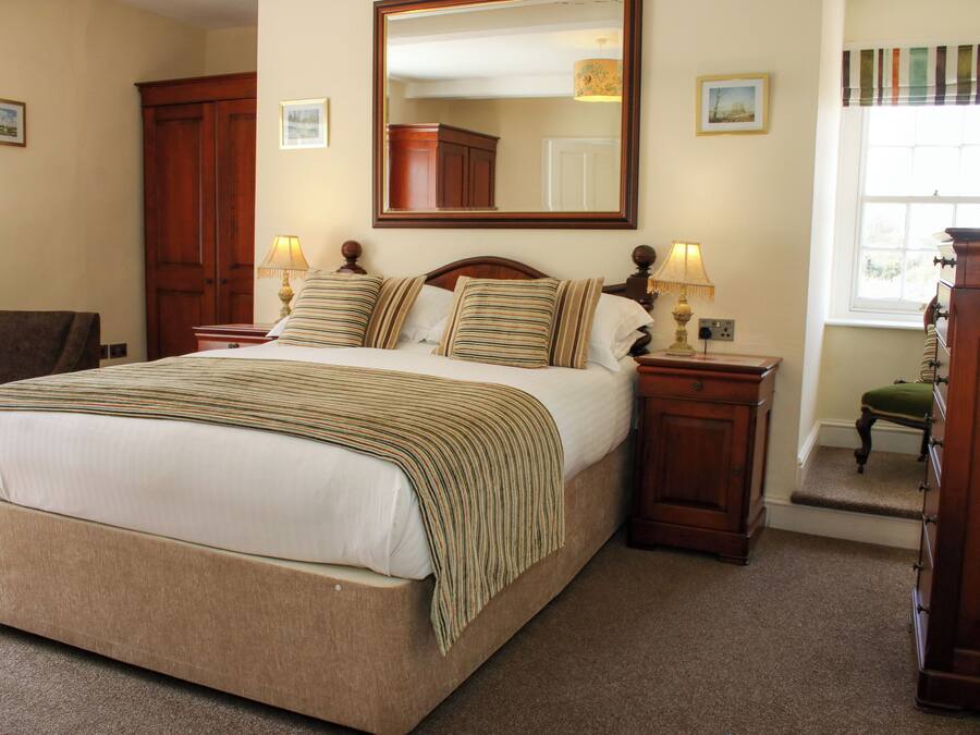 A bedroom with a large bed, wooden headboard, two bedside tables with lamps, a mirror above the bed, a wooden wardrobe, and a window with a view.