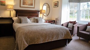 Una habitación de hotel con una cama grande, un rincón de descanso con dos sillones, un aparador de madera, un espejo y una ventana con vistas al exterior.