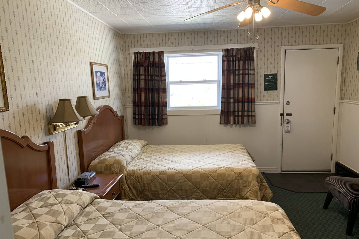 A hotel room with two beds, a ceiling fan, a window with curtains, a bedside table with a lamp, and a door with a sign.