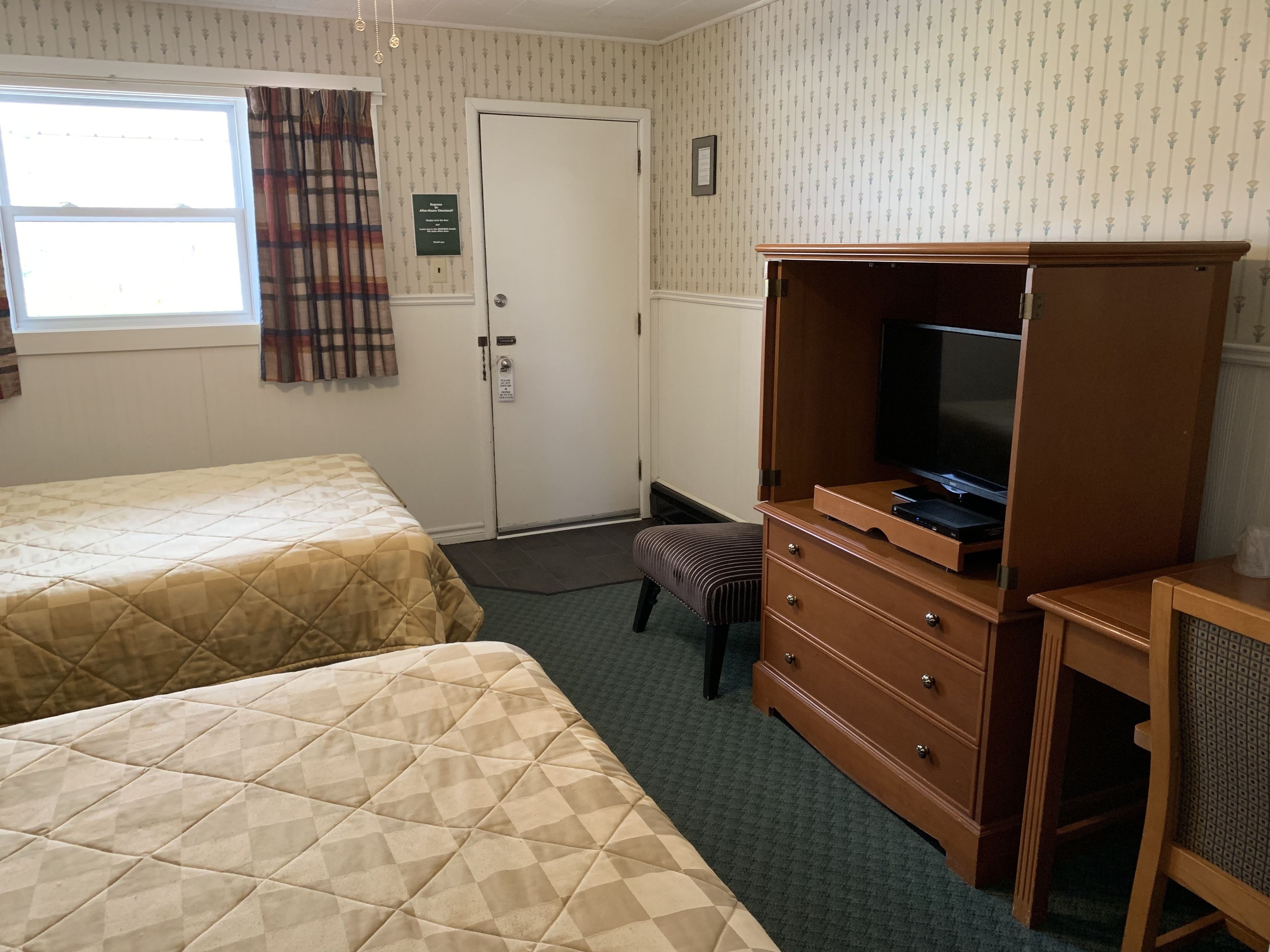 A hotel room with two beds, a wooden dresser with a television, a door with a sign, and a window with curtains.
