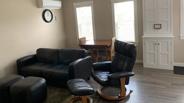 A living room with a black leather sofa, a black leather armchair, and a wooden coffee table. There is a dining table with chairs, a clock on the wall, and a window with blinds. - The Lionstone Inn Motel and Cottages (Pictou)