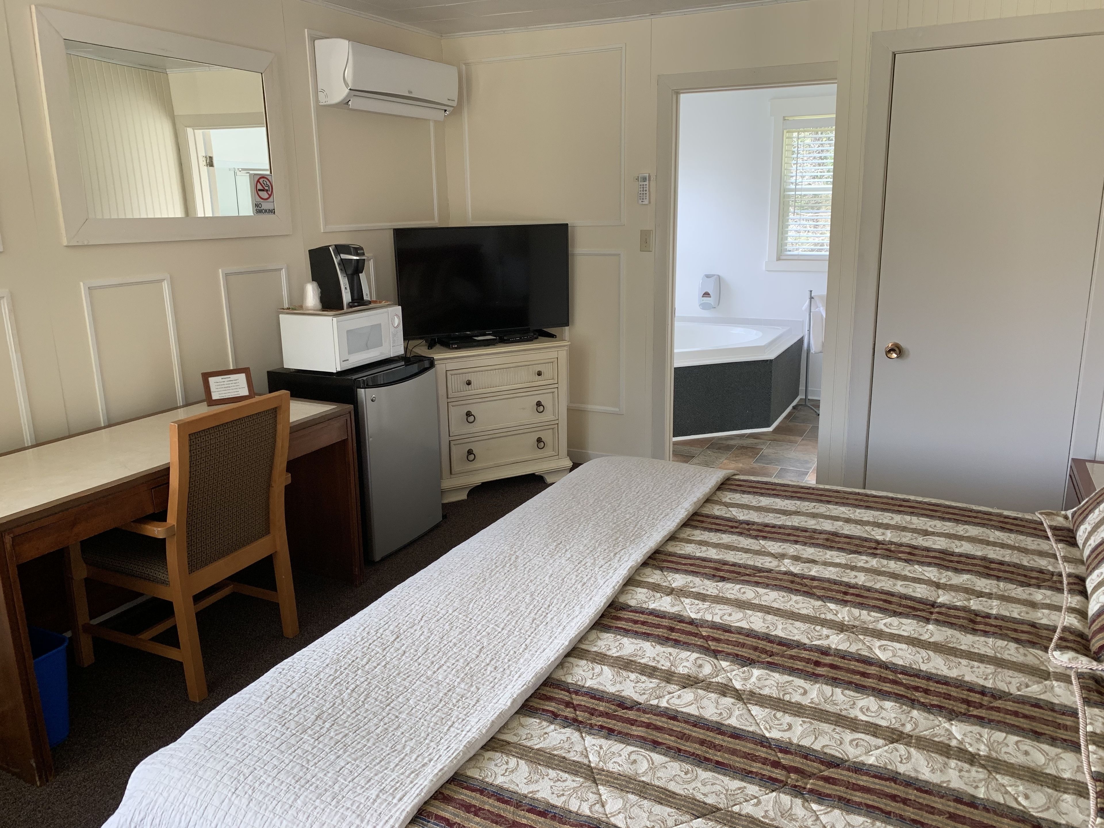 A hotel room with a bed, a desk with a chair, a television, a microwave, a small refrigerator, a bathroom with a bathtub, and a door.