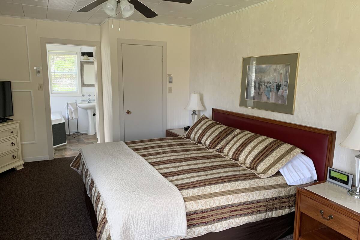 A bedroom with a bed, a bedside table with a lamp, a ceiling fan, a bathroom with a sink and mirror, and a door leading to another room.