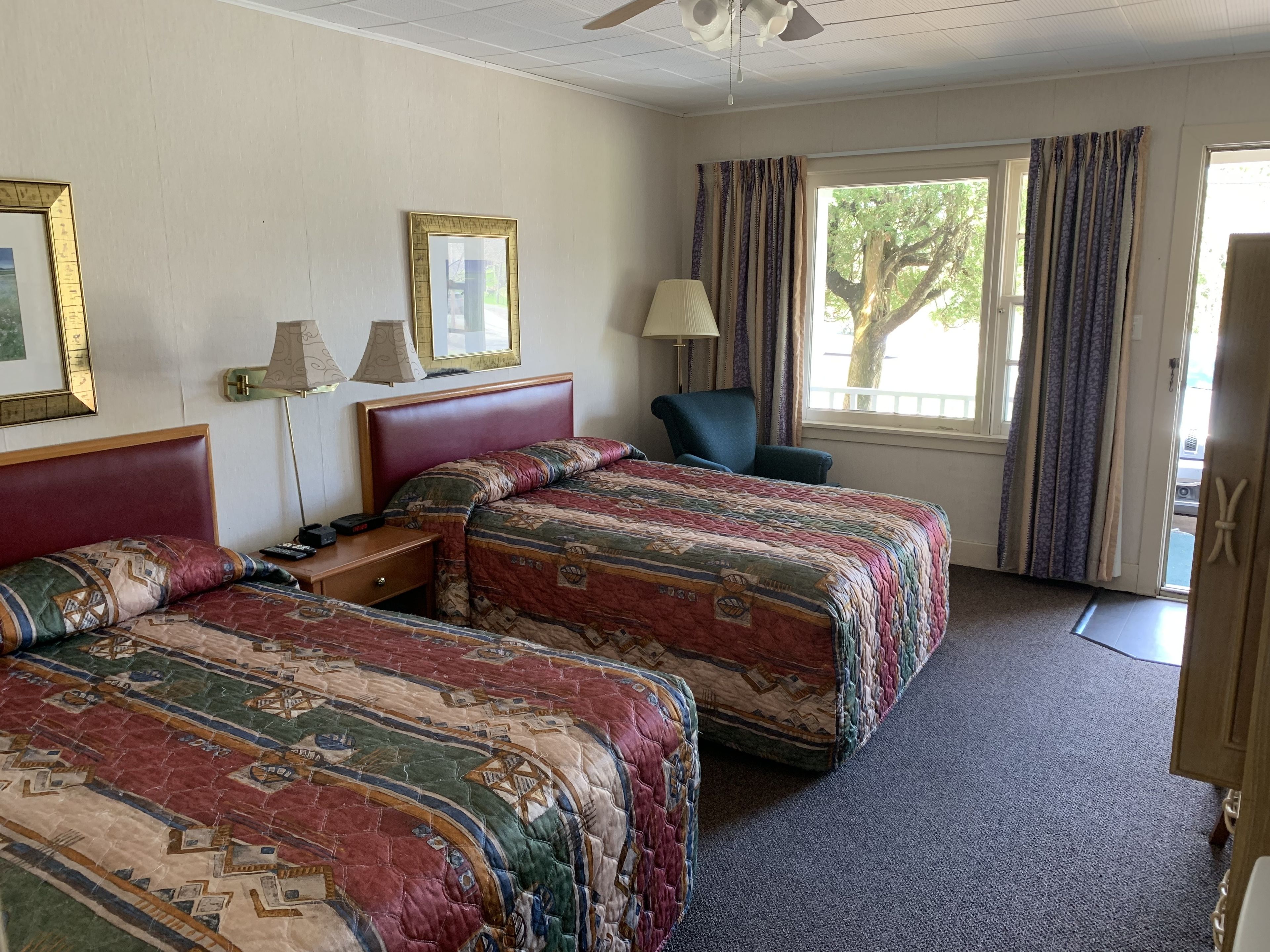 A hotel room with two beds, a desk, a chair, a window with curtains, and a ceiling fan.
