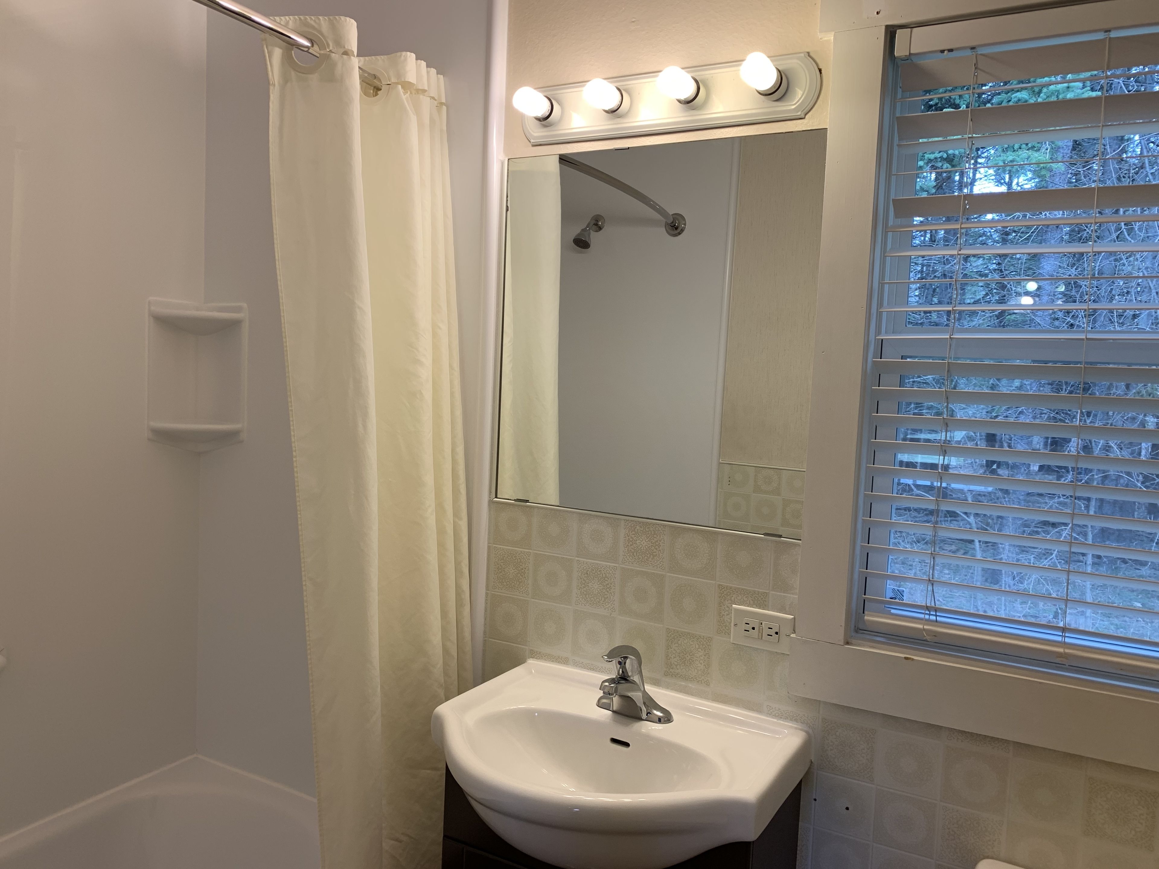 Un baño con lavamanos blanco, un espejo con luces, cortina de ducha y una ventana con persianas.