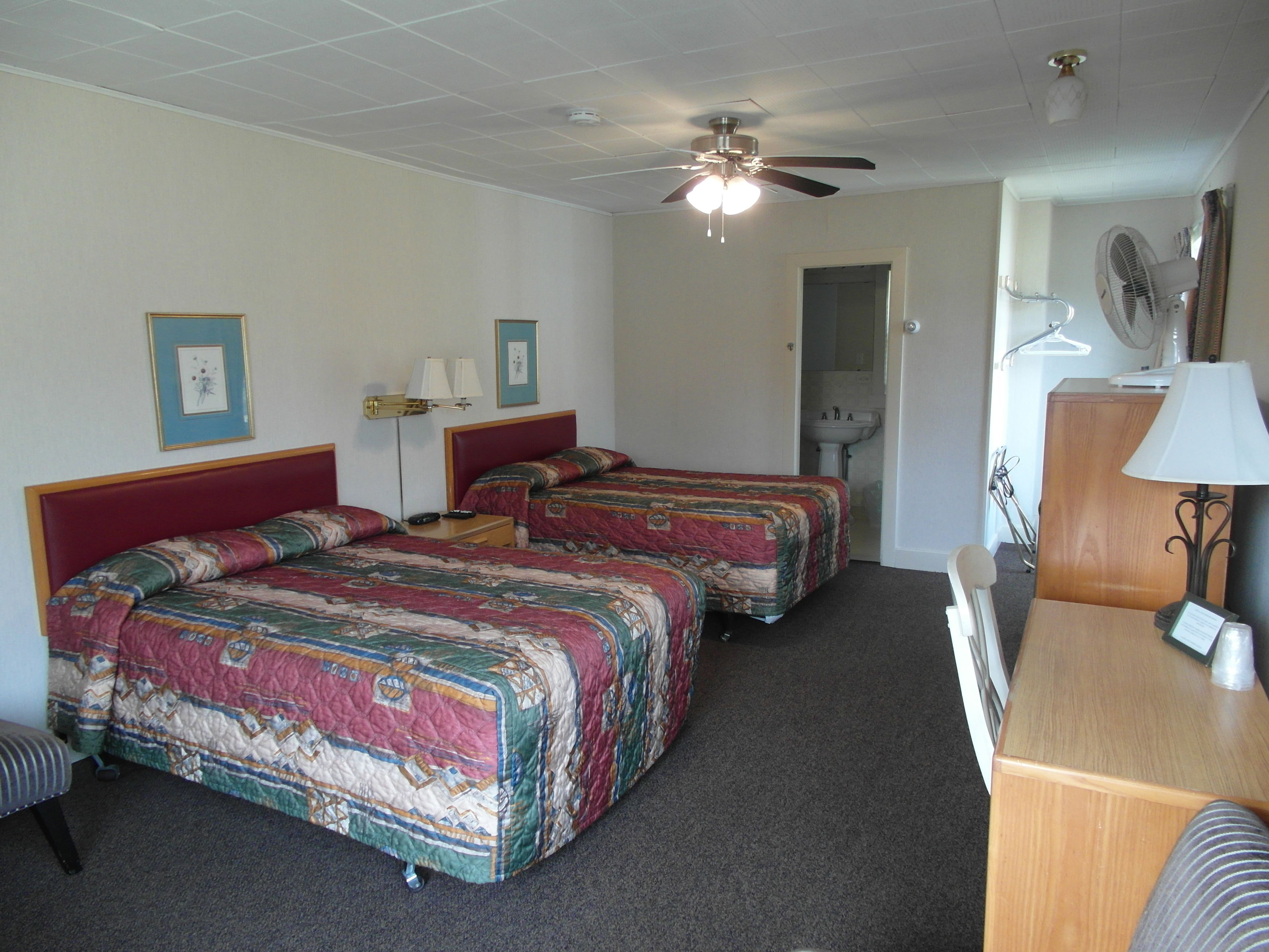 A hotel room with two beds, a ceiling fan, a desk with a lamp, and a mirror.