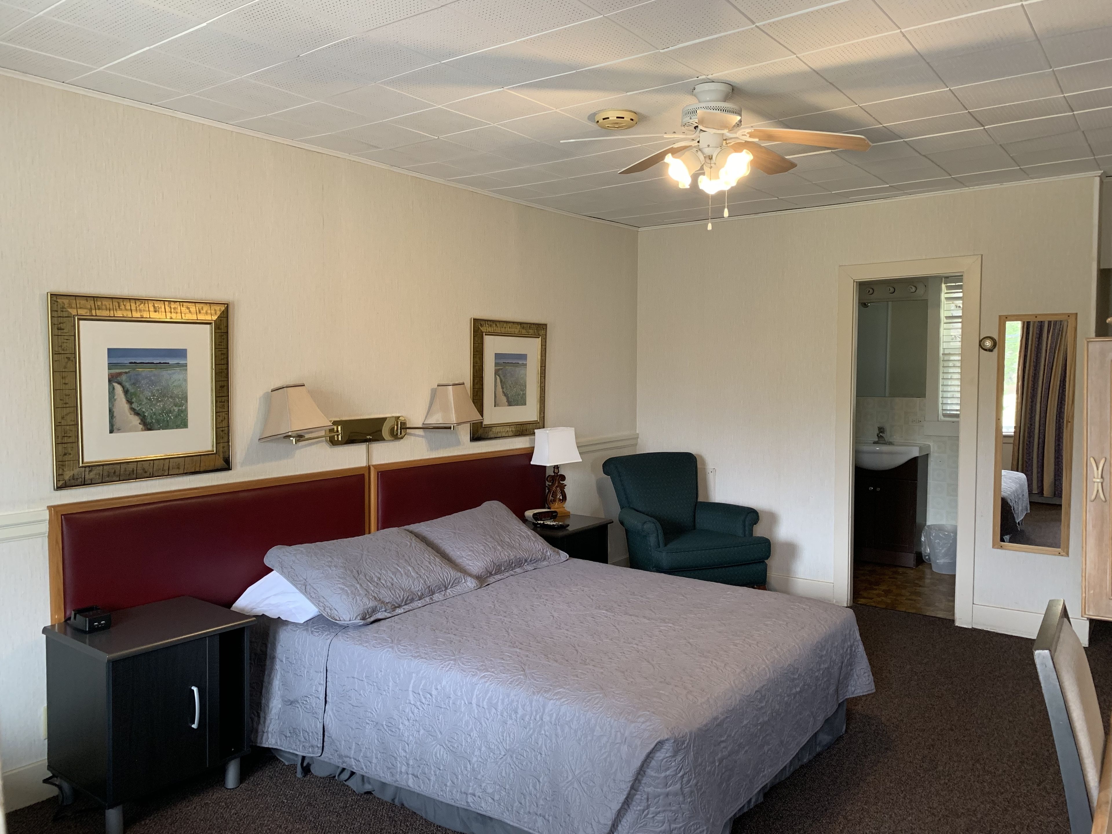 A hotel room with a bed, a nightstand, a chair, a desk, a mirror, and a bathroom visible through an open door.