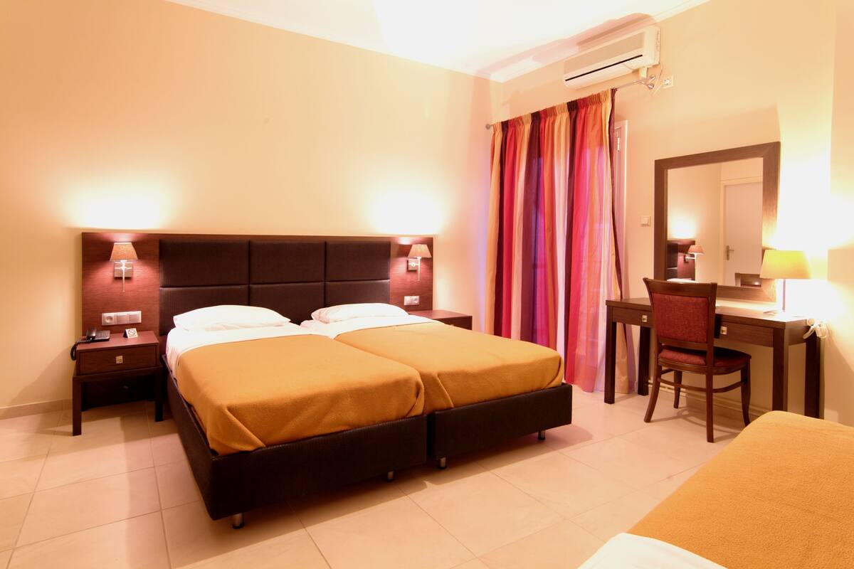 A hotel room with a double bed, a desk with a chair, a nightstand, and an air conditioning unit.