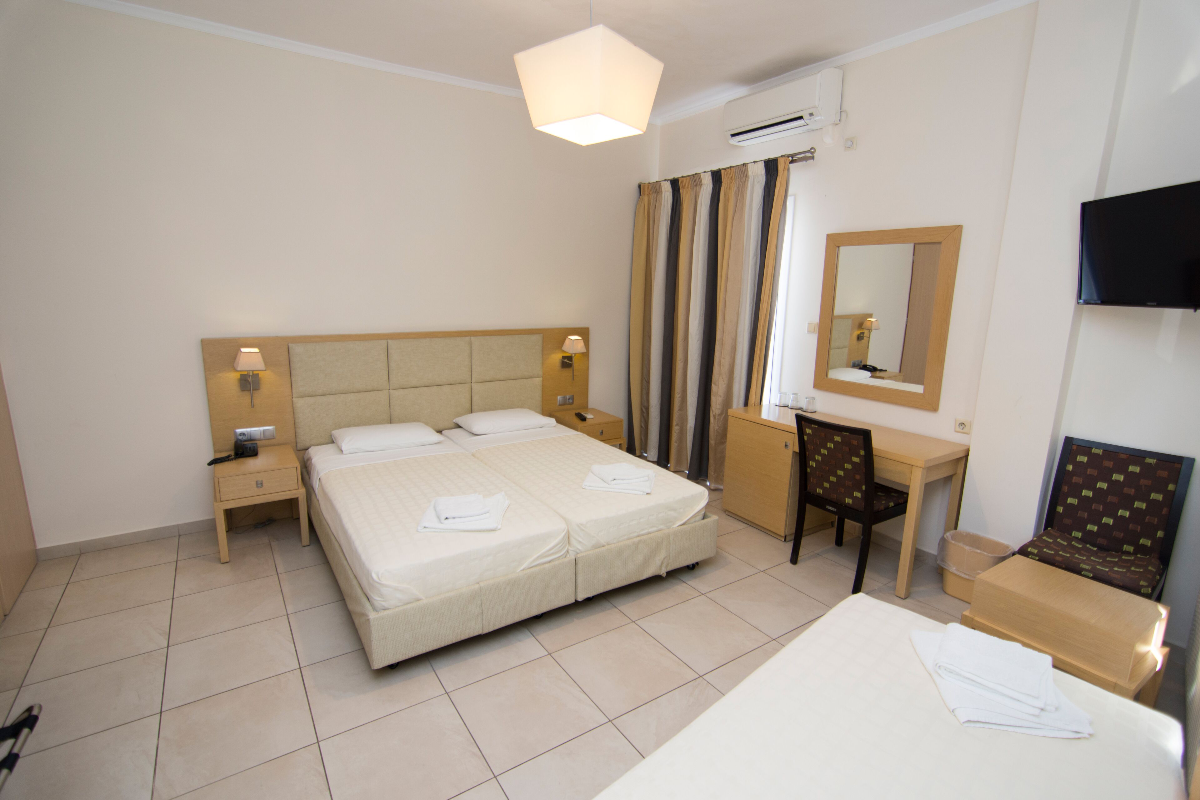 A hotel room with a large bed, bedside tables, a desk, a chair, a TV, and an air conditioning unit.