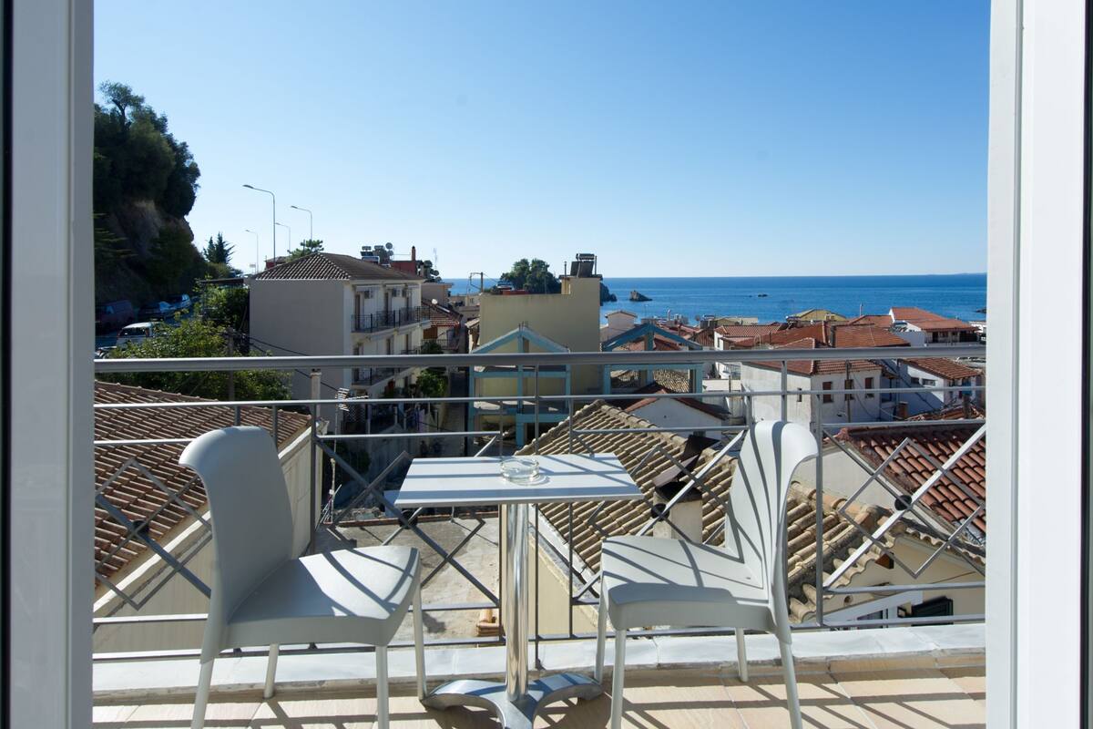 A balcony with a table and chairs, overlooking a coastal town and the sea.