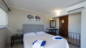Quarto de hotel com uma cama grande, mesa de cabeceira, escrivaninha, cadeira, espelho e janela com cortinas.