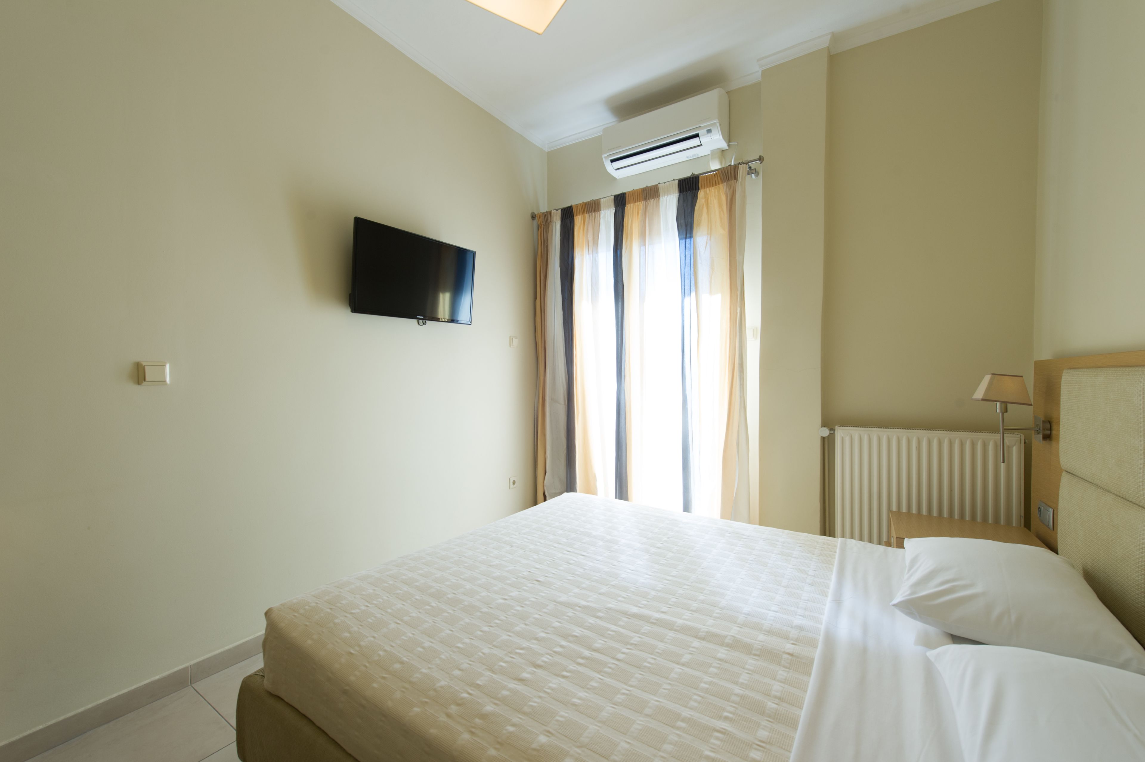 Habitación de hotel con cama, televisión montada en la pared, equipo de aire acondicionado, ventana con cortinas y mesita de noche con lámpara.