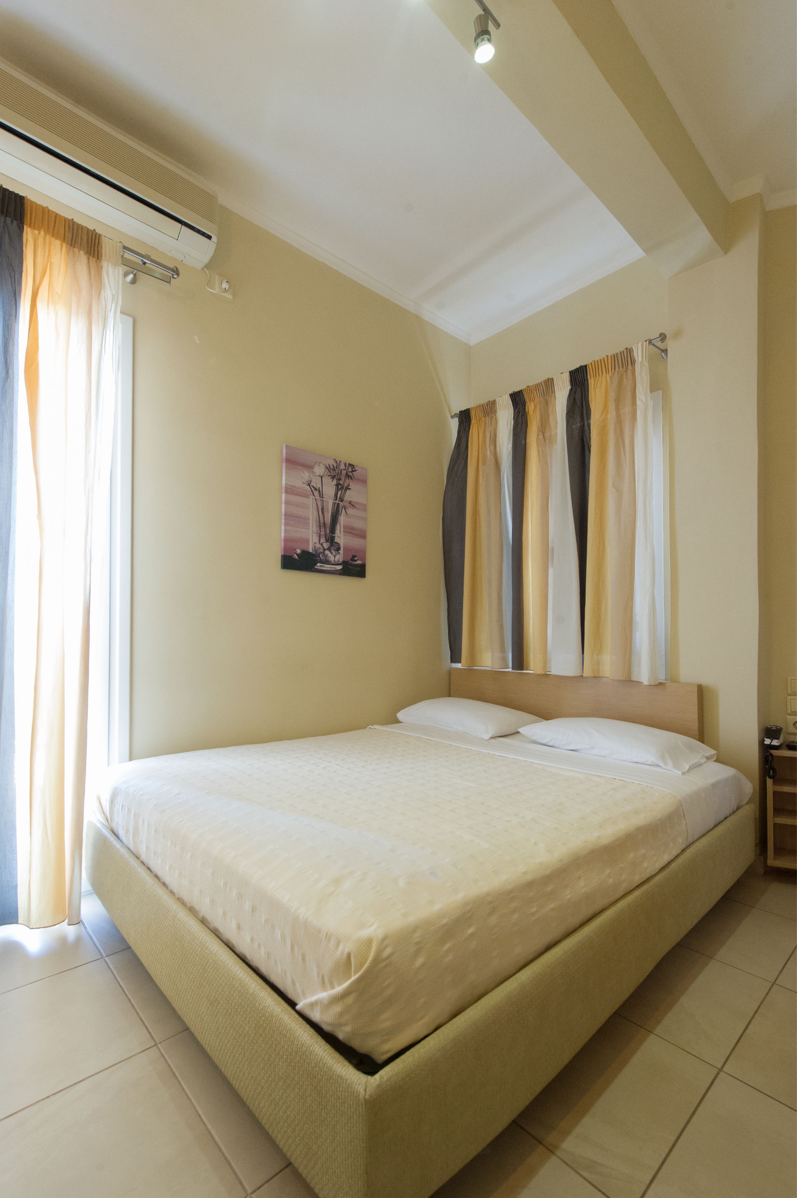 A hotel room with a bed, air conditioning unit, curtains, and a painting on the wall.
