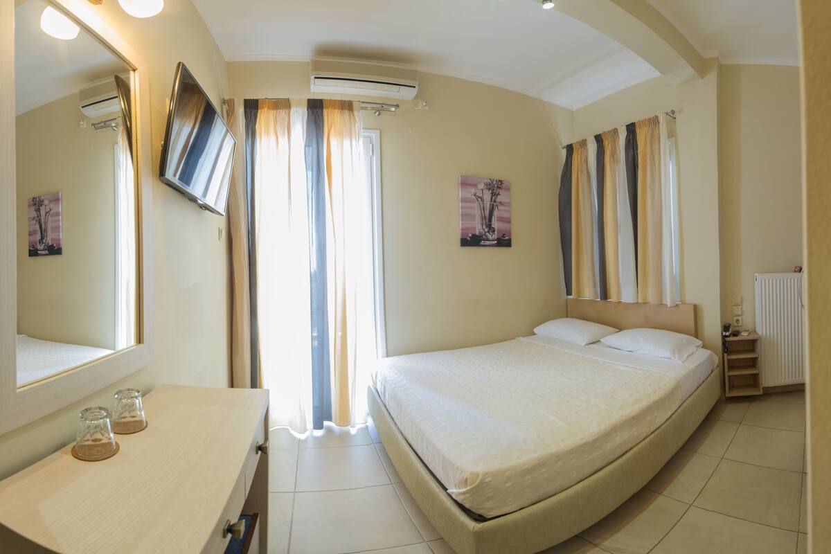 A modern hotel room with a large bed, a flat-screen TV, a desk with two glasses, and a window with curtains.