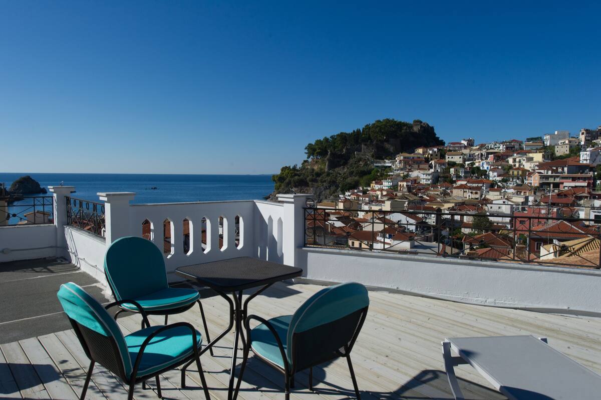 A terrace with a table and chairs, overlooking a coastal town with buildings and a clear sky.