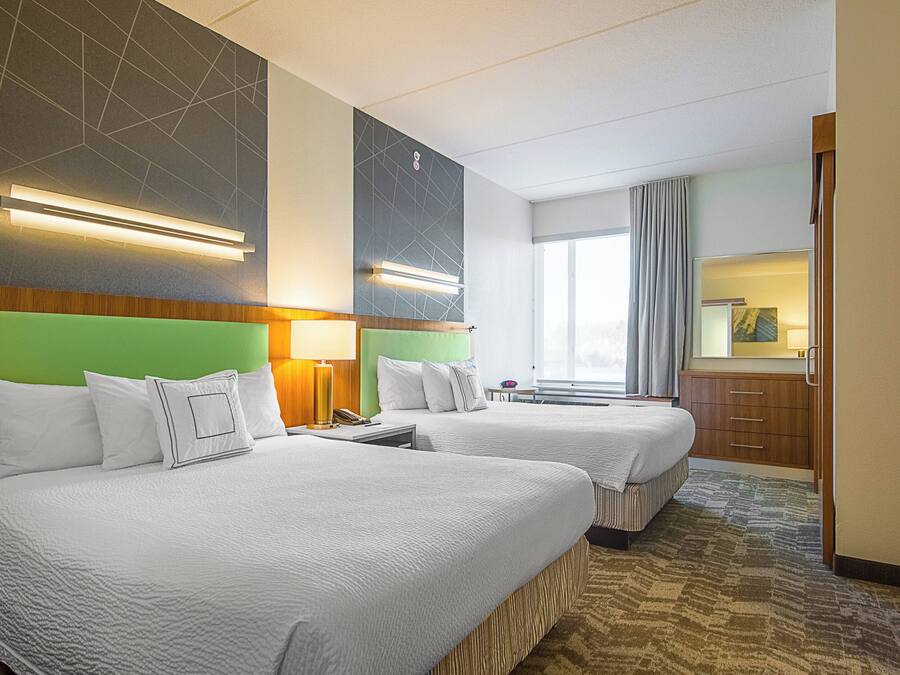 A hotel room with two beds, a large window with curtains, a wooden cabinet, and a wall-mounted light fixture.