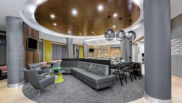 A modern lounge area with a curved ceiling, grey sofas, and a central dining table with chairs. - SpringHill Suites by Marriott San Antonio SeaWorld Lackland (San Antonio)