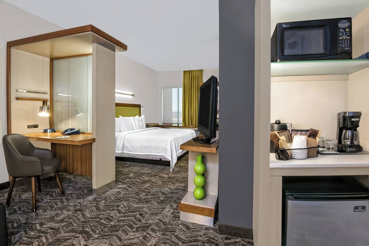 A hotel room with a bed, desk, chair, TV, microwave, coffee maker, and refrigerator.