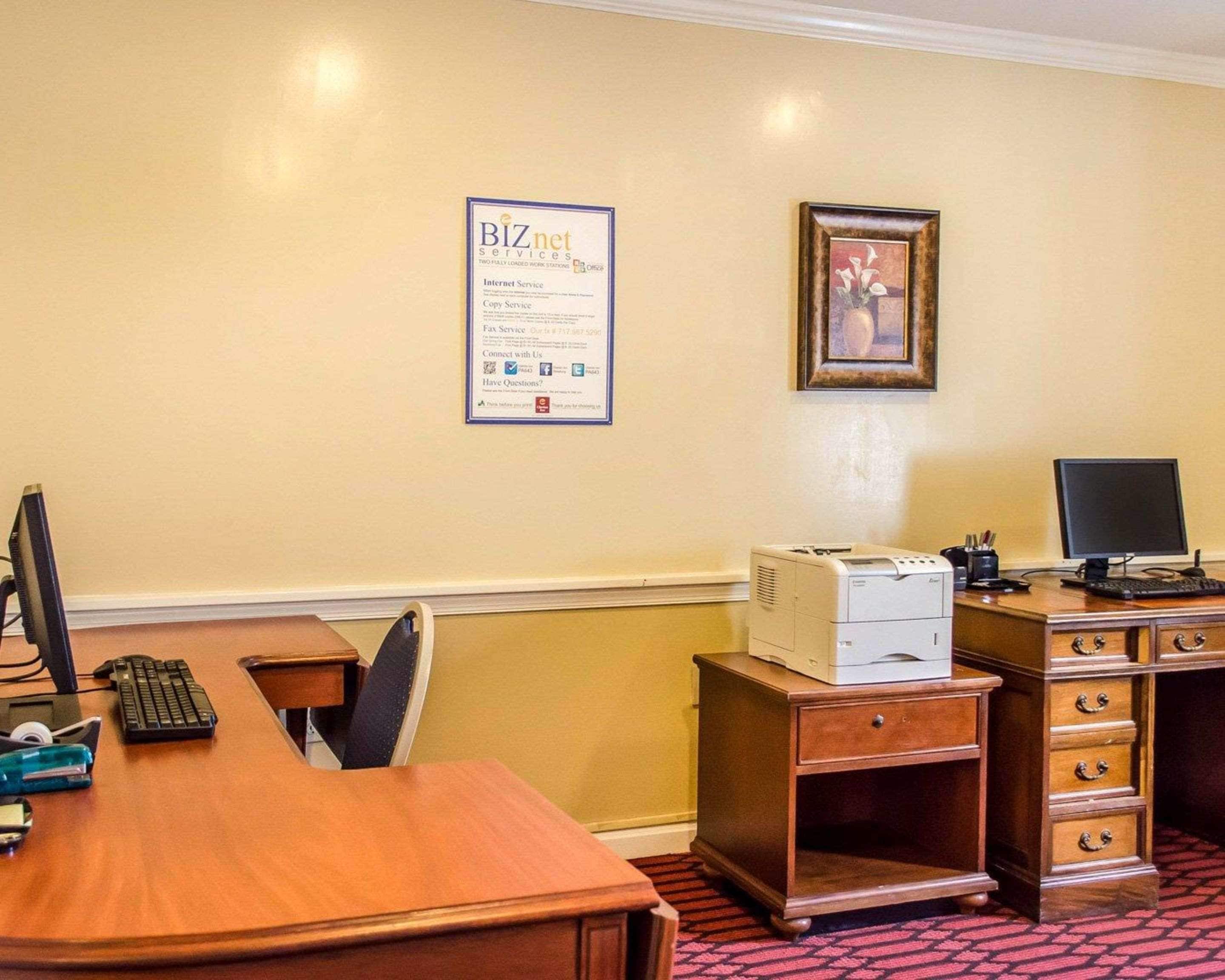 A hotel room with a wooden desk, a computer, a printer, and a framed picture on the wall.