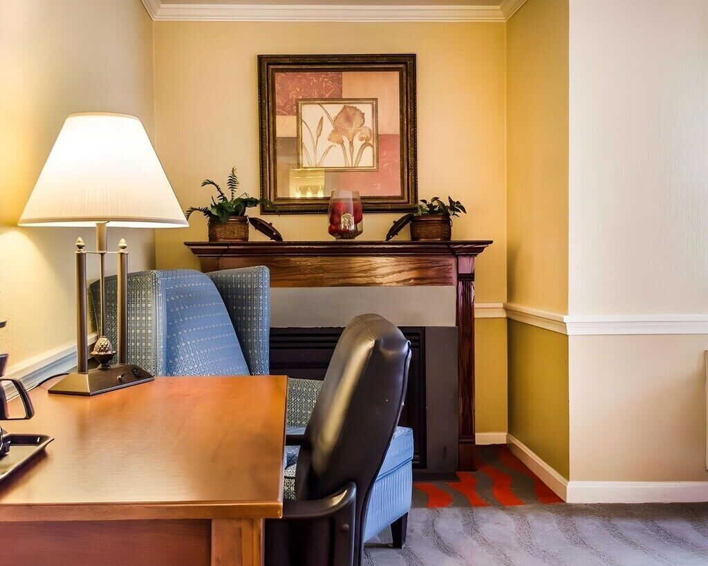 A room with a wooden desk, a chair, a lamp, a fireplace, and a framed picture on the wall.
