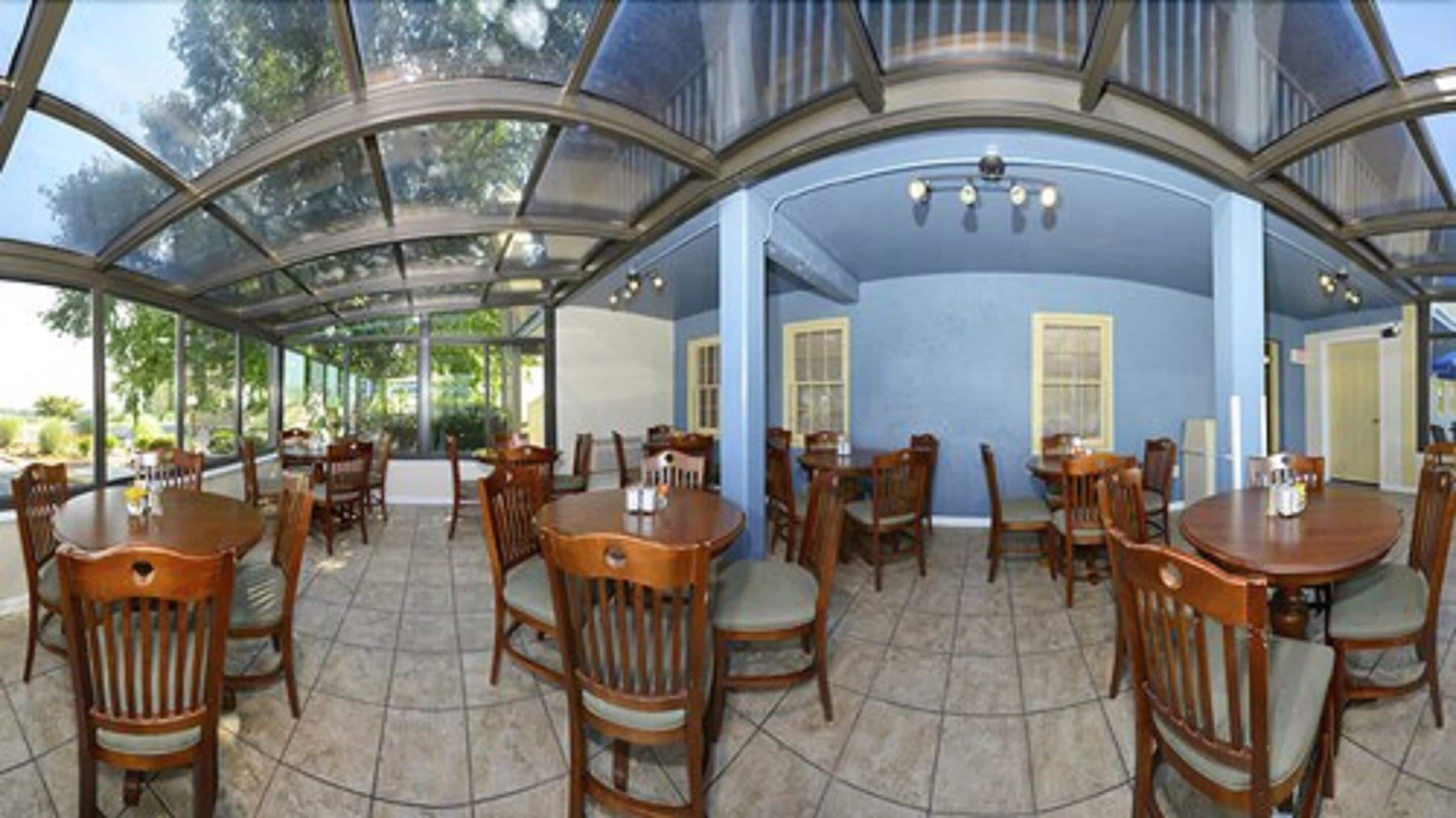Un espace extérieur abrité, aménagé pour les repas, avec plusieurs tables et chaises en bois, de grandes fenêtres et un sol carrelé.
