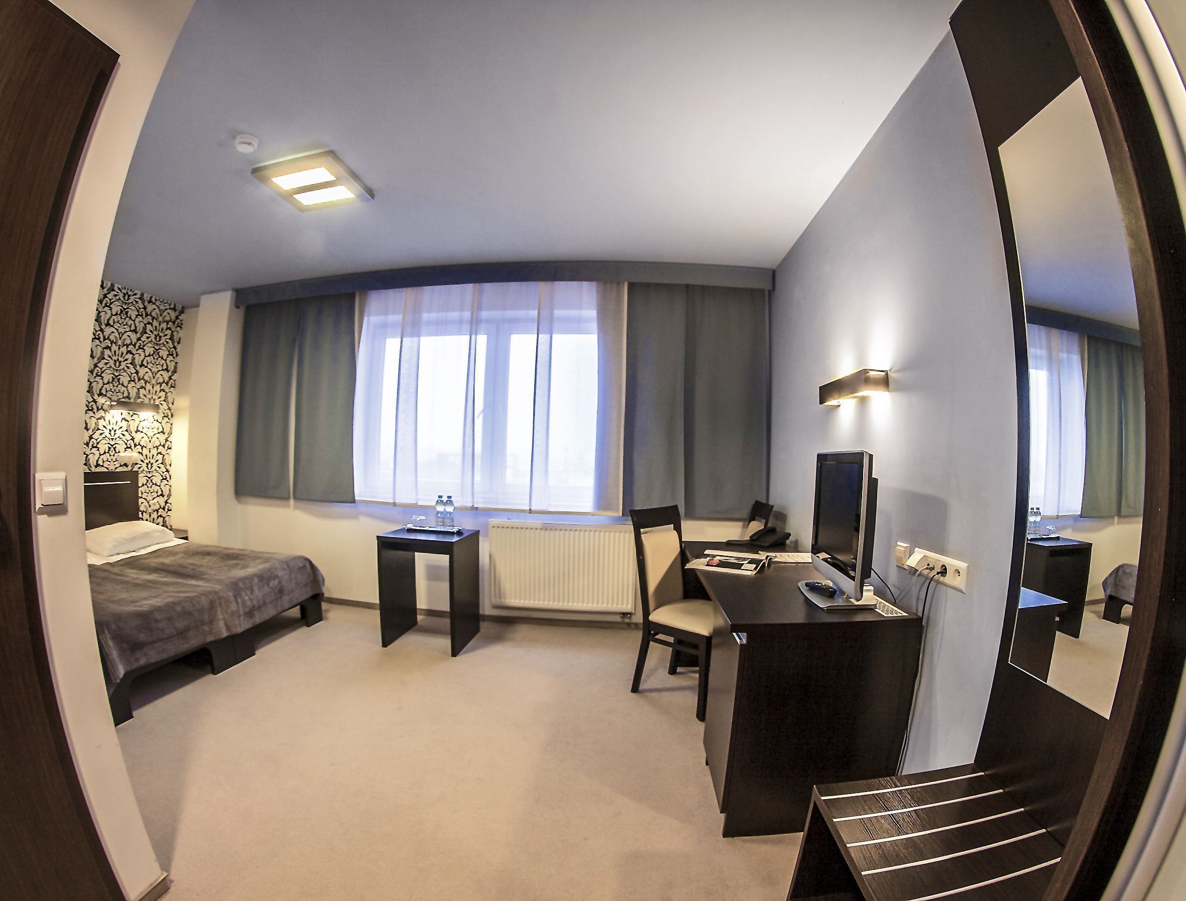 A compact hotel room with a bed, a desk, a chair, a computer, and a large mirror.