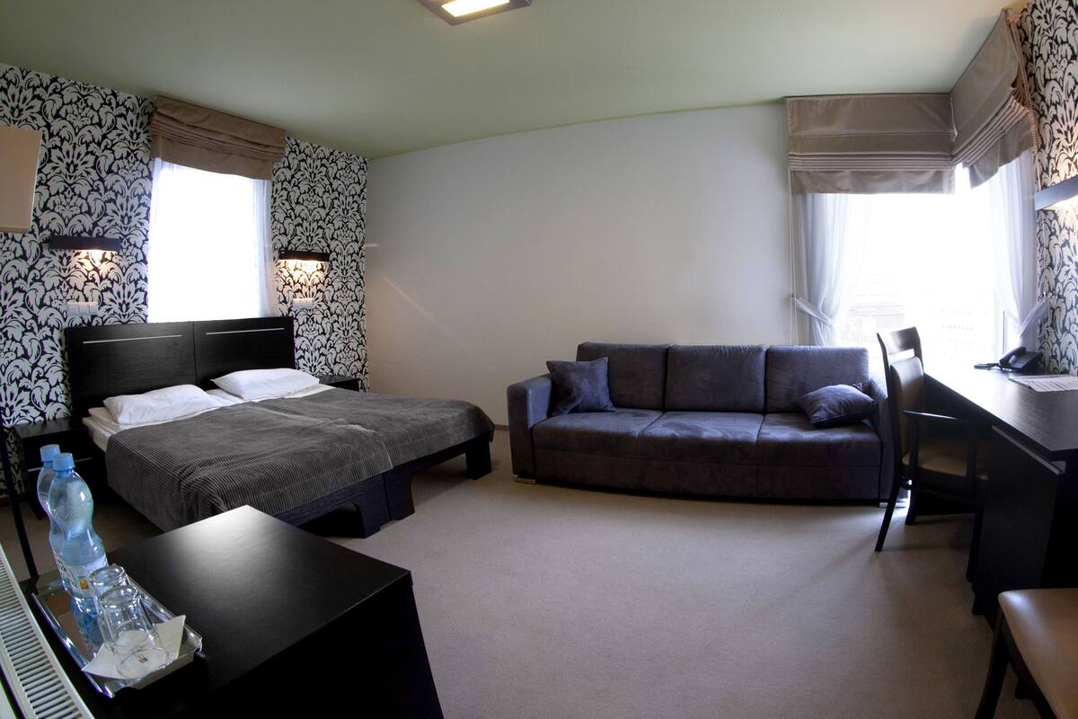 A hotel room with a bed, a sofa, a desk, and a chair. There are two windows with curtains, a wall with a patterned wallpaper, and a ceiling light.