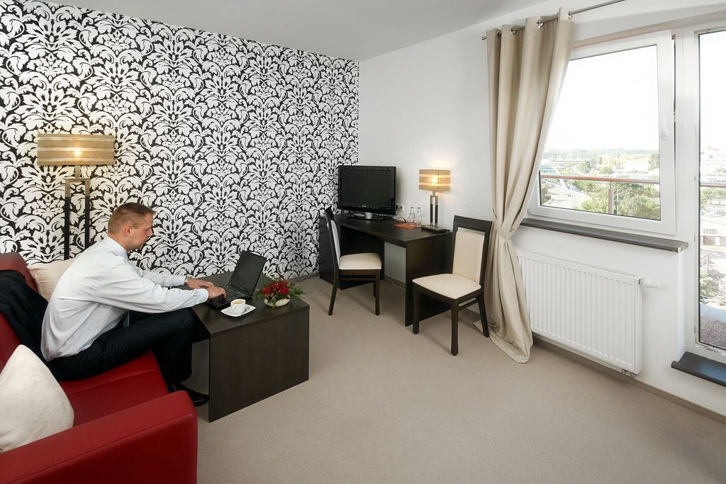 Habitación de hotel con un sofá rojo, un escritorio con computadora, un televisor y una ventana con cortinas.