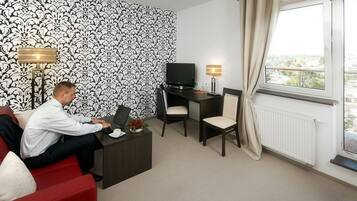 Habitación de hotel con un sofá rojo, un escritorio con computadora, un televisor y una ventana con cortinas.