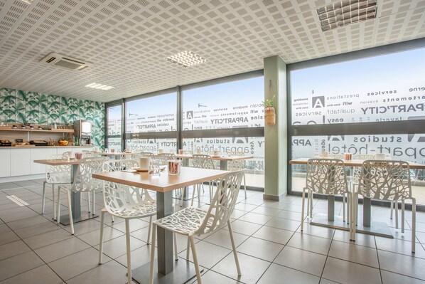A modern dining area with white chairs and a wooden table, large windows with a view, and a ceiling with a geometric pattern. - Appart'City Classic Nîmes Centre (Nîmes)