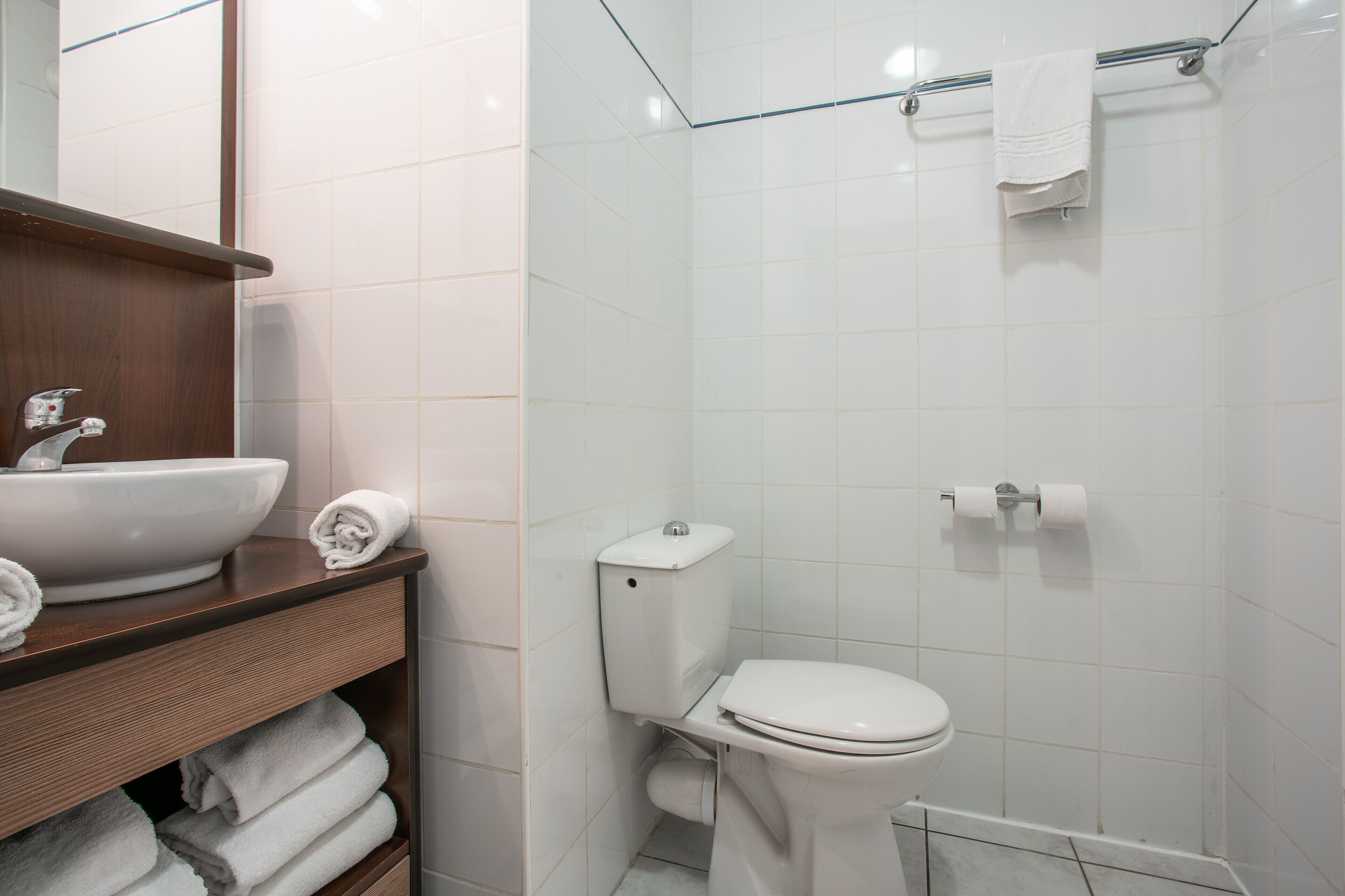 Un baño con lavamanos, inodoro y ducha blancos. Hay toallas en la encimera y un perchero sobre la ducha.