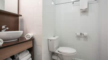 Une salle de bain avec un lavabo, des toilettes et une douche, le tout de couleur blanche. On trouve des serviettes sur le comptoir et un porte-serviettes au-dessus de la douche.