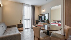 A hotel room with a sofa, a dining table with chairs, a TV, a bed with a red bedspread, and a sliding door leading to another room. - Appart'City Classic Nîmes Centre (Nîmes)