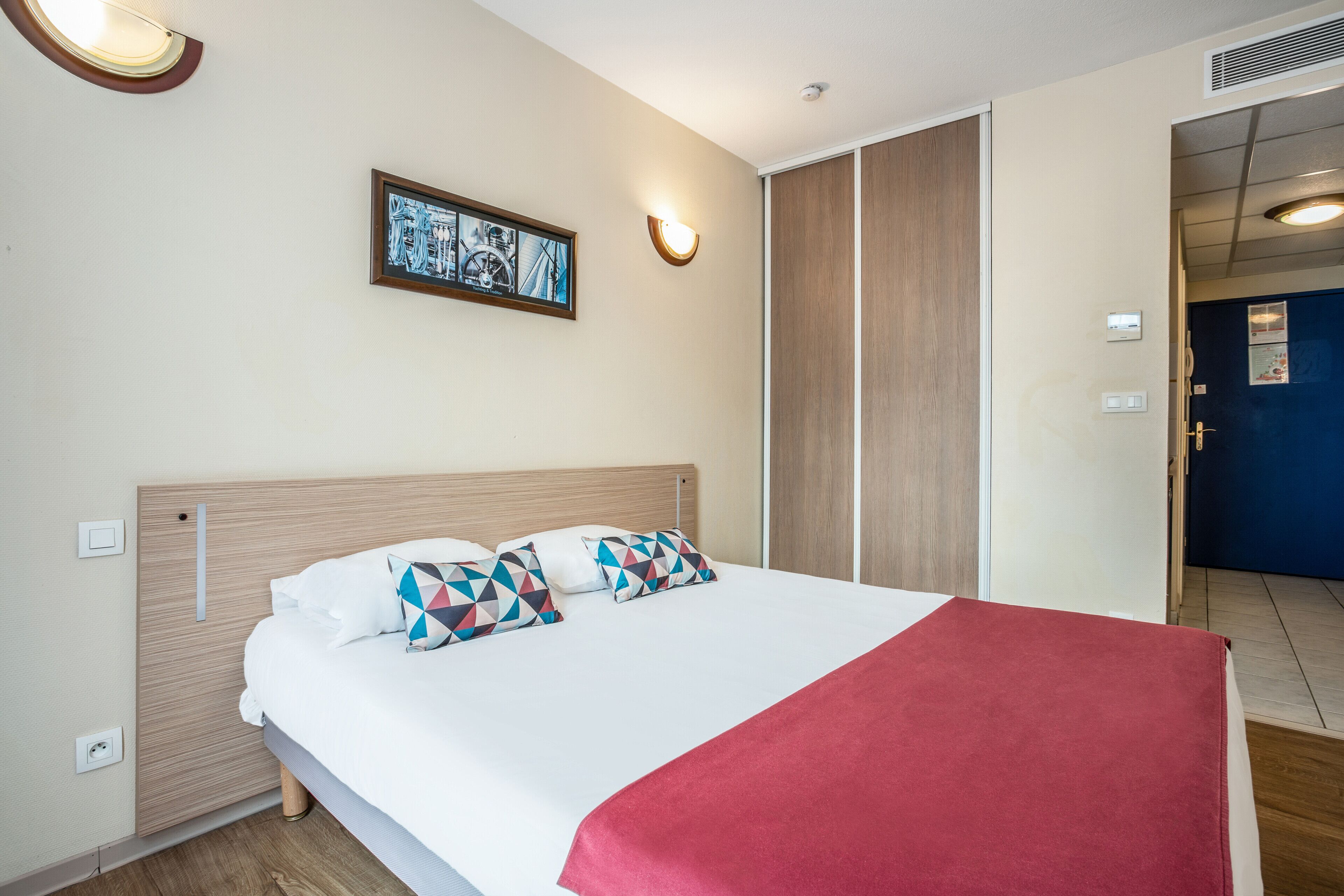 A hotel room with a bed, a wall-mounted picture, a light fixture, and a door.
