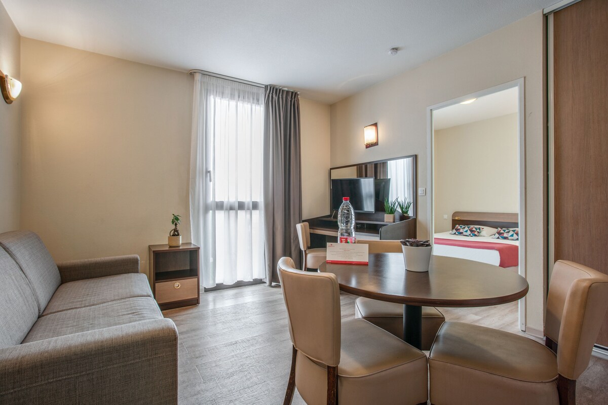 A hotel room with a sofa, a dining table with chairs, a TV, a bed with a red bedspread, and a sliding door leading to another room.