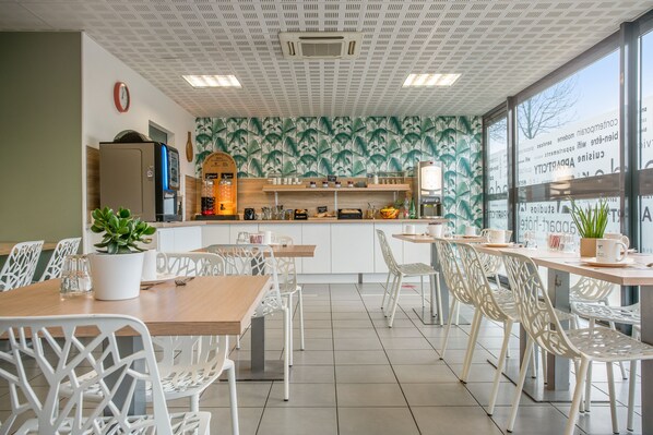 Una cafetería moderna con sillas blancas, mesas de madera, una zona de cocina y un amplio ventanal con vistas.