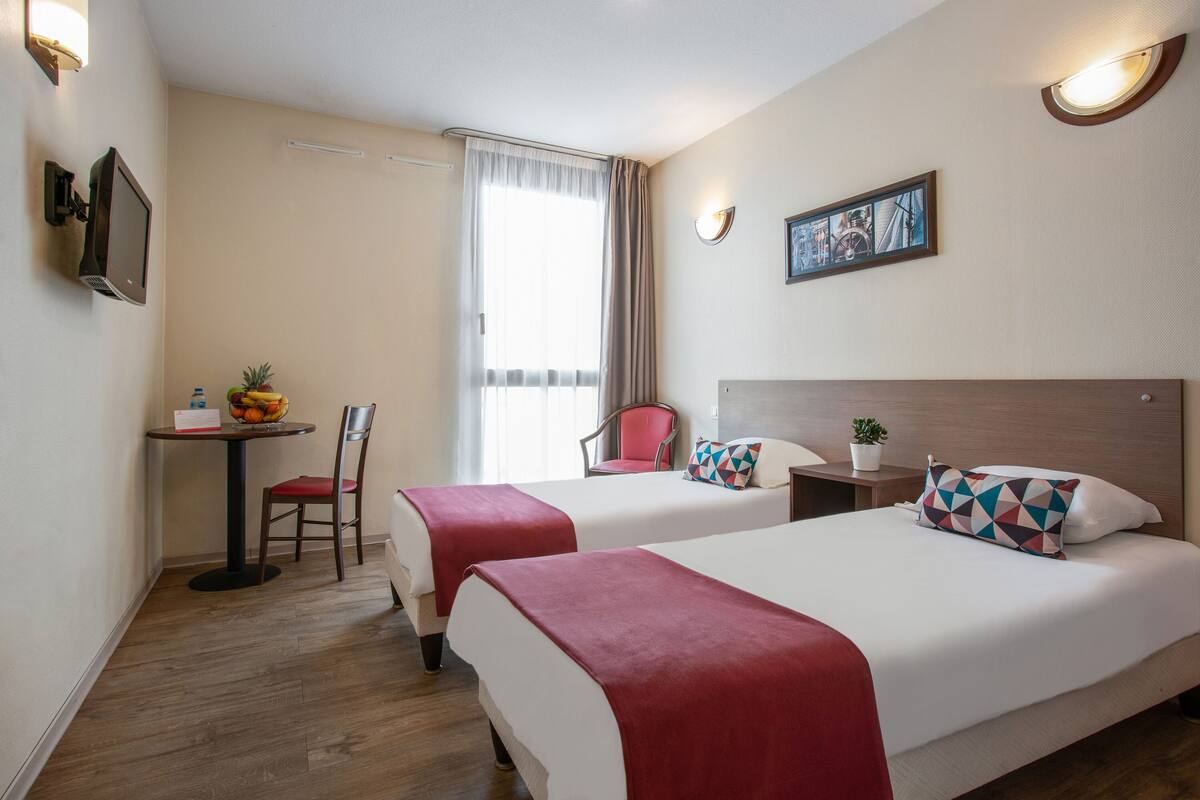 A hotel room with two beds, a desk with a chair, a small table with fruit, and a window with curtains.