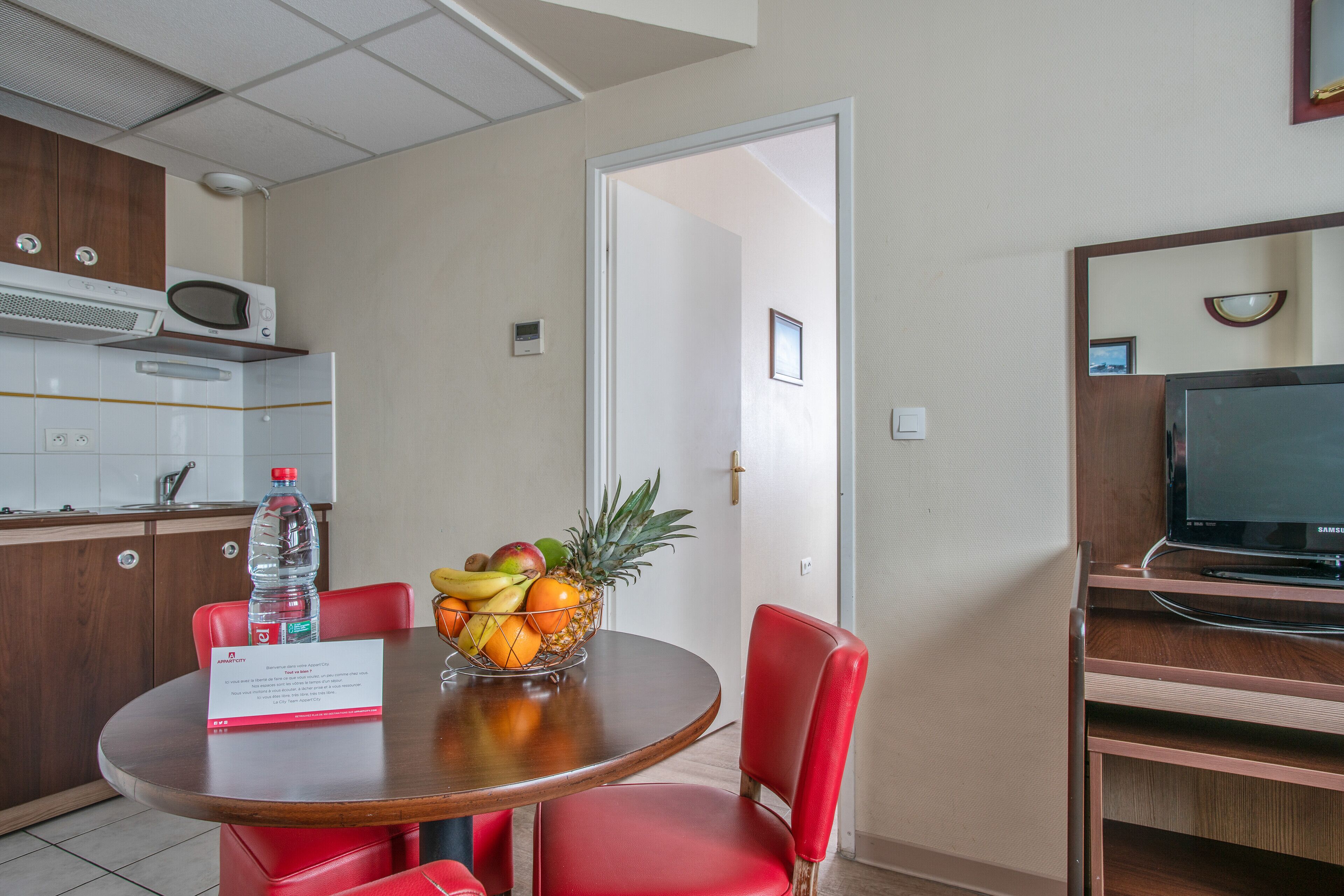 Habitación de hotel con comedor que incluye una mesa redonda, sillas rojas y un frutero.