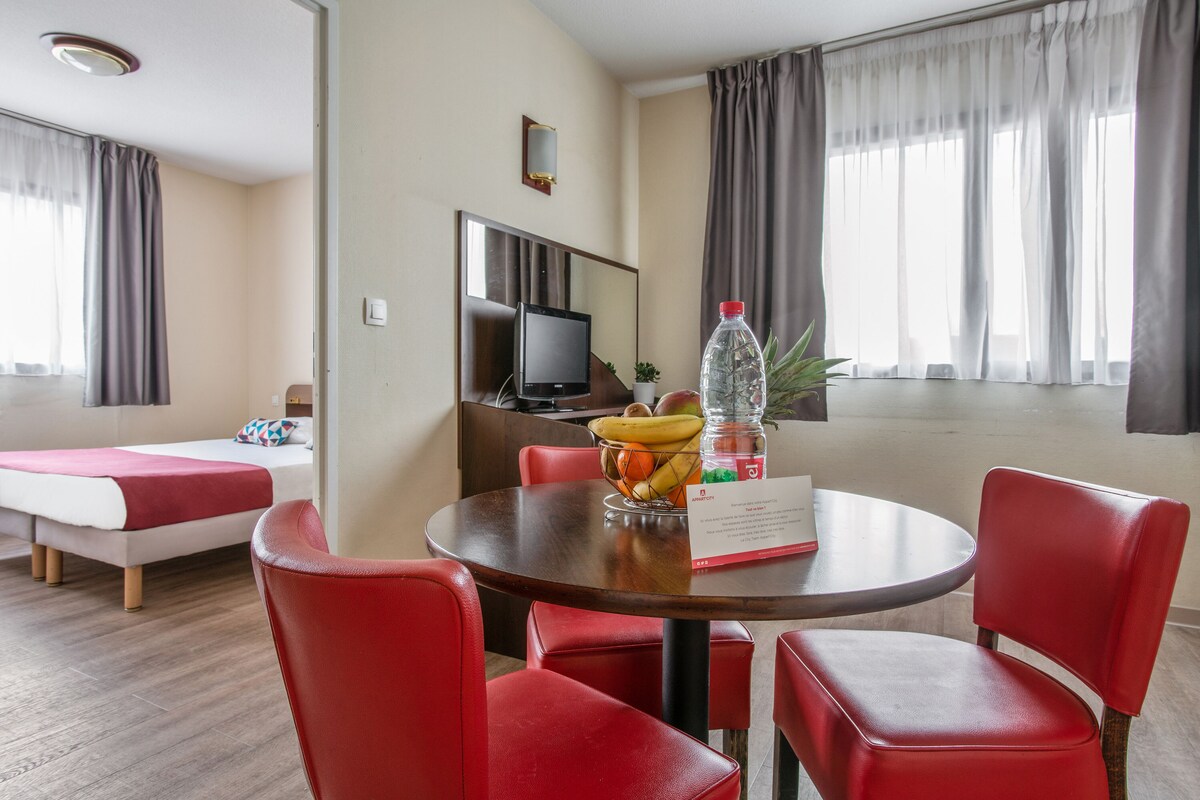 A hotel room with a bed, a dining table with red chairs, a TV, and a window with curtains.