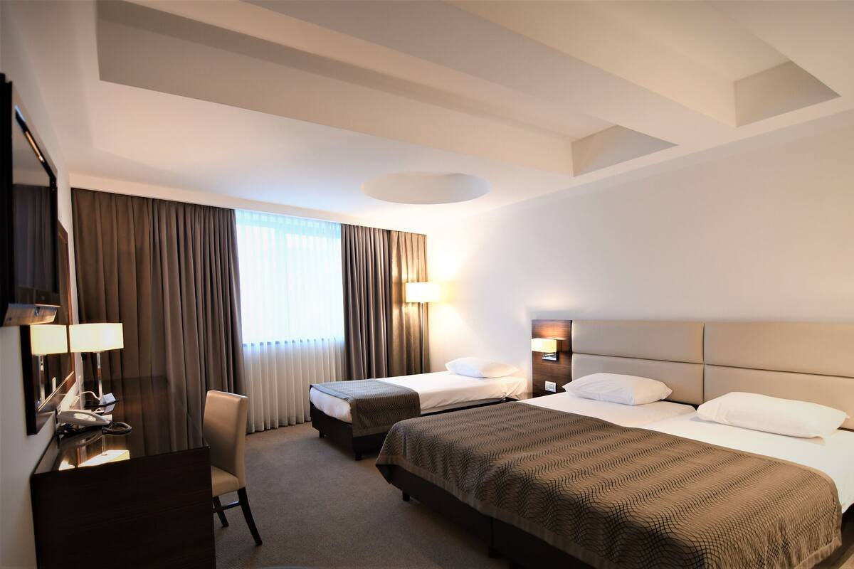 A hotel room with two beds, a desk, a chair, a lamp, and a large window with curtains.