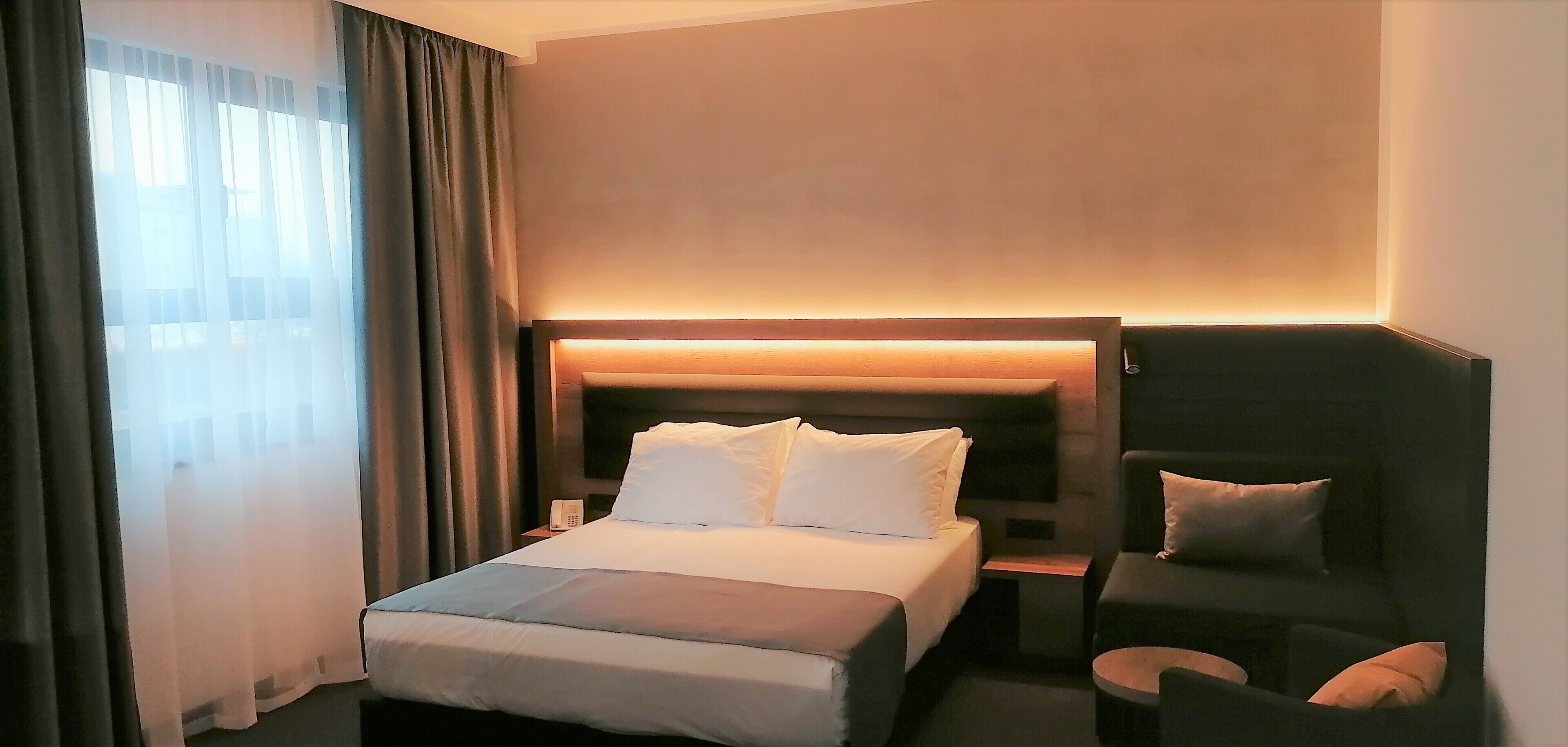 A hotel room with a bed, a bedside table, a chair, and a headboard with built-in lighting.