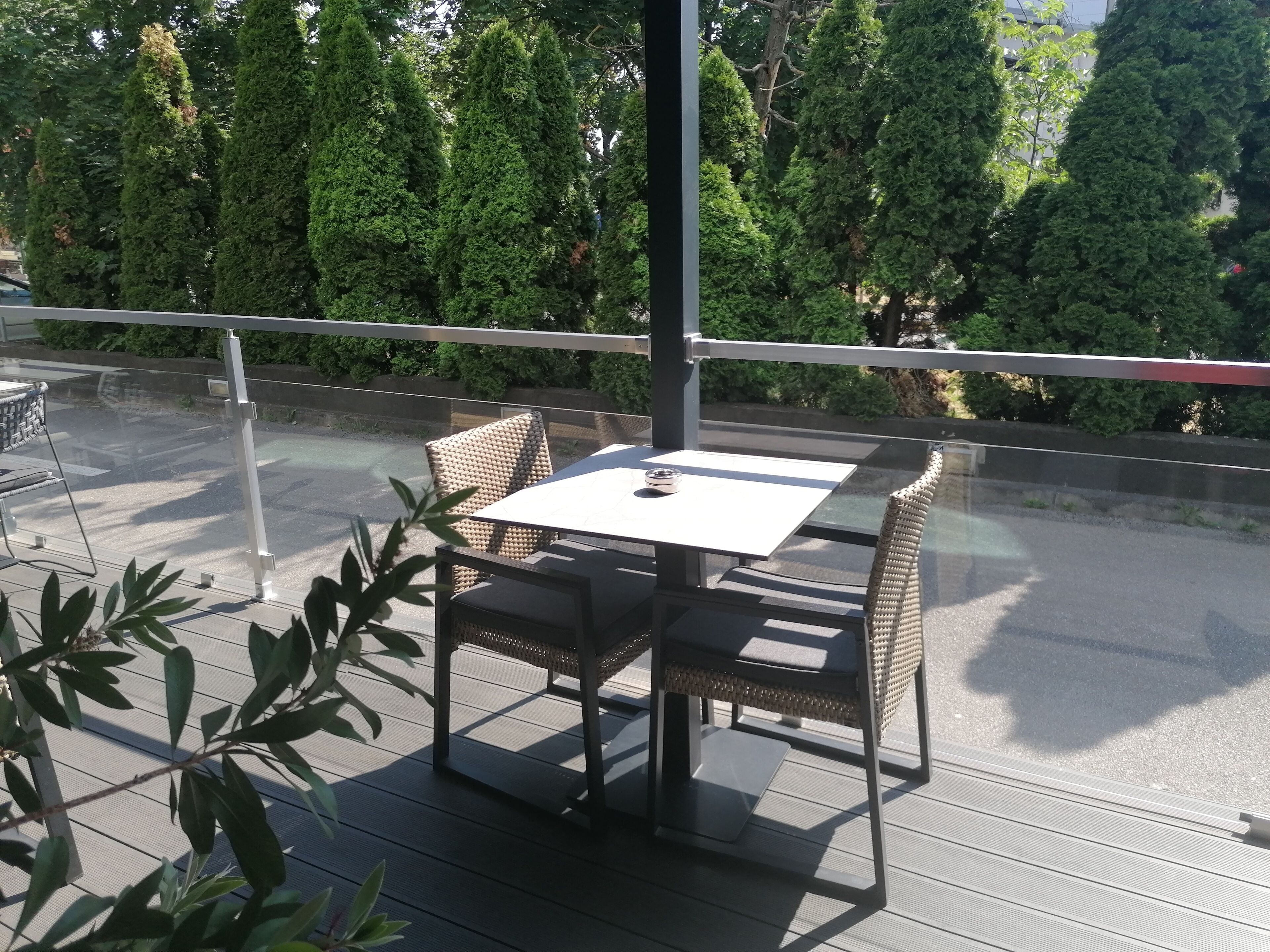 An outdoor seating area with a table and chairs, surrounded by a railing, overlooking a road and greenery.