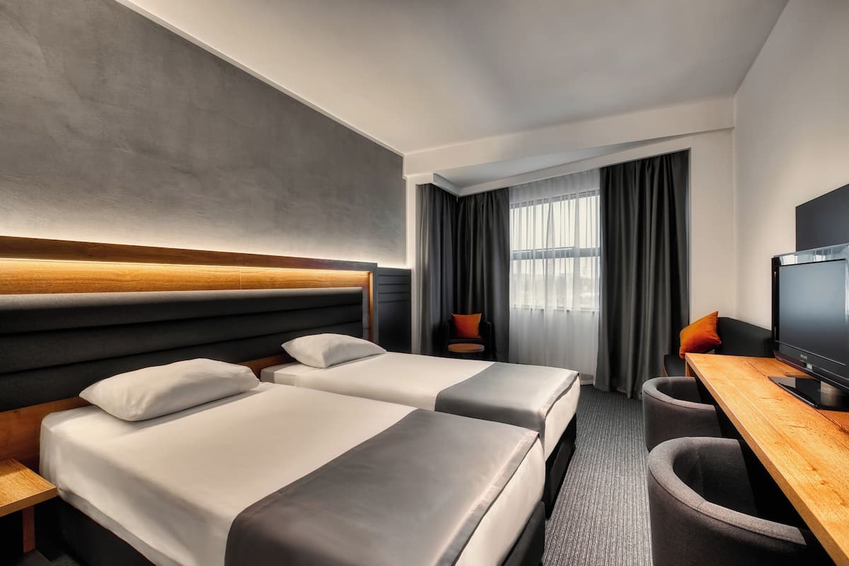 A hotel room with two beds, a desk with a television, and a view of the outdoors through the window.