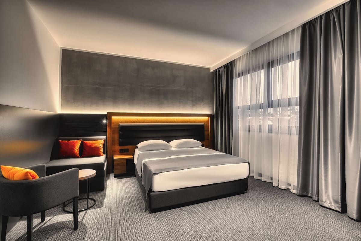 A modern hotel room with a large bed, a sofa, a small table, and a headboard with built-in lighting.