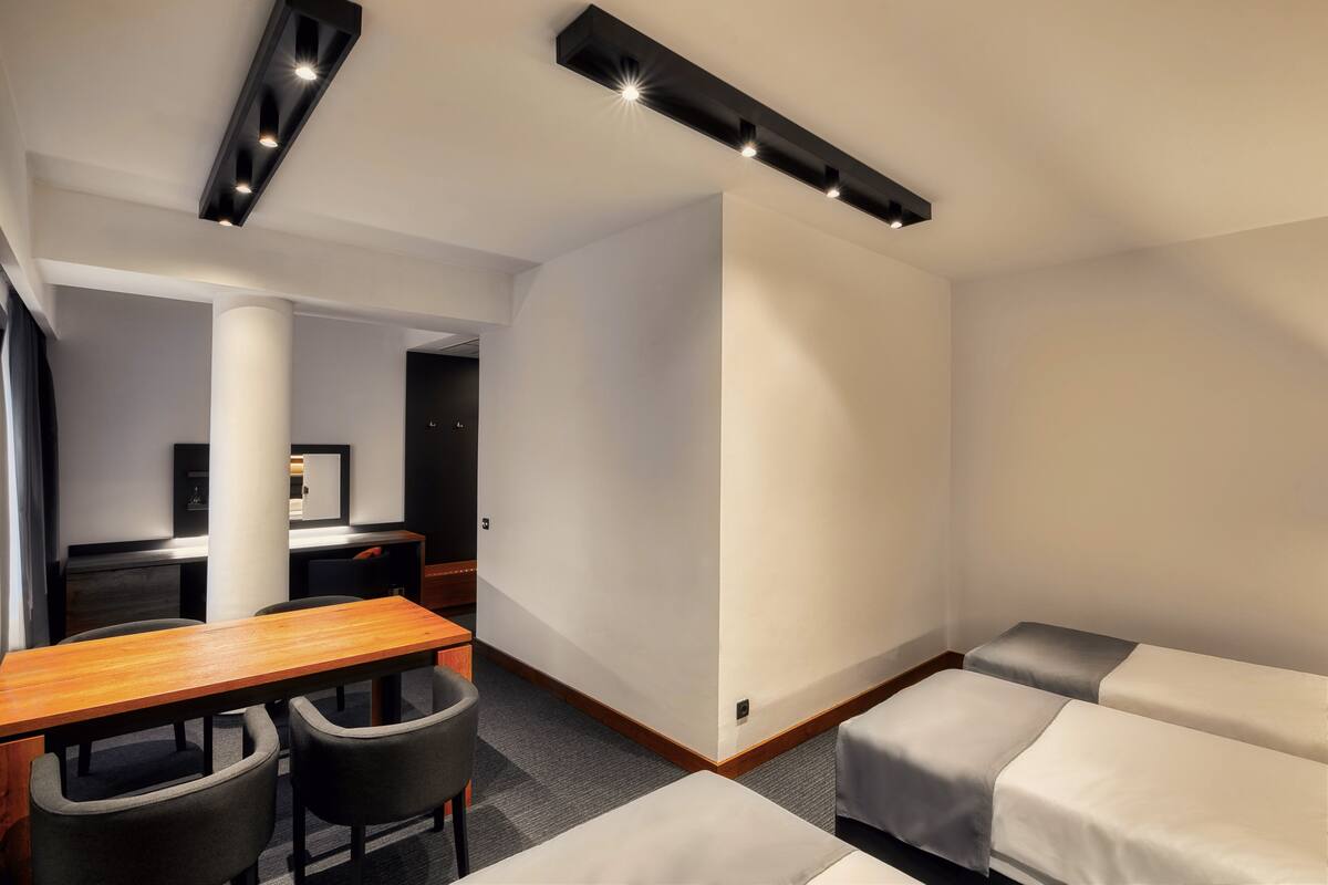 A modern hotel room with a wooden table, two gray chairs, a desk, and two single beds.